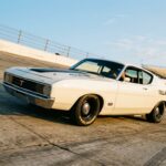 A 1969 Ford Torino Talladega in Wimbledon White photographed at low angle on a sunlit banked oval superspeedway, the reshaped tapered nose and long sloping Sportsroof fastback silhouette prominently visible, dramatic perspective emphasizing the car's aerodynamic profile against a vast expanse of concrete banking and pale blue sky, cinematic editorial automotive photography with golden hour lighting casting long shadows, photorealistic detail capturing the clean flush bodywork and understated muscle car stance, evoking purposeful speed and precision engineering rather than theatrical flamboyance