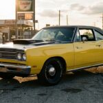 A 1969 Plymouth Road Runner in vivid yellow with a black hood scoop sits on a gritty American street at golden hour, photographed from a low three-quarter front angle. The car's muscular B-body lines are sharply defined in the warm late-day light, with chrome details catching the sun and slightly worn asphalt beneath the wide tires suggesting real-world use rather than a showroom. The setting is working-class Americana — a gas station and chain-link fence blurred softly in the background, telephone poles lining the road. The mood is unpretentious and powerful, celebrating raw mechanical purpose over luxury. The image has a cinematic, editorial quality with rich film-like colors, deep shadows under the wheel arches, and an atmosphere of authentic 1960s American muscle culture — fast, honest, and built for the street.