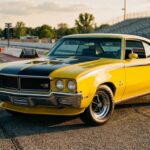 A 1970 Buick GSX Stage 1 in bold Saturn Yellow with black racing stripes, parked on a sunlit dragstrip apron, shot from a low three-quarter front angle that emphasizes the muscular hood with its tachometer, front spoiler, and aggressive stance. The car sits alone, commanding the frame, with a blurred dragstrip and grandstand in the soft background. The lighting is warm and golden, late afternoon sun raking across the sheet metal to reveal every line and curve of the body. The overall mood is confident and understated menace — a car that looks fast without trying too hard, refined yet coiled with latent power. Photorealistic editorial style, cinematic depth of field, rich color saturation, clean composition with no text or logos visible.