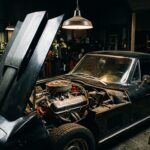 A dramatic, cinematic close-up photograph of a 1967 Chevrolet Corvette C2 Sting Ray in a dark, moody garage setting, illuminated by a single overhead industrial light casting sharp shadows. The car is finished in a deep, aggressive color — dark blue or black — with the long hood dominating the foreground. The hood is propped open, revealing a massive 427 cubic inch big block engine with a single four-barrel Holley carburetor and open-element air cleaner, chrome and cast iron gleaming under the focused light. The fastback roofline and twin round taillights are visible in the background. The scene feels raw, purposeful, and stripped-down — no radio, no frills, just a machine built entirely around performance. Shot in an editorial automotive photography style with high contrast, shallow depth of field, and a gritty, documentary realism that conveys the car's uncompromising racing DNA and mythological rarity.