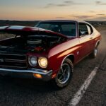 A dramatic, cinematic close-up shot of a 1970 Chevrolet Chevelle SS 454 in a deep, bold color — either Cranberry Red or Fathom Green — photographed at dusk on an empty American highway stretching into the horizon. The car sits low and aggressive with its long hood dominating the frame, blacked-out grille, hood scoop, and SS badging visible. Warm golden hour light rakes across the muscular body lines and factory rally wheels, casting long shadows on the asphalt. The engine bay hood is slightly raised, hinting at the massive big-block V8 beneath. The atmosphere is cinematic and reverent, evoking raw American mechanical power at its absolute peak — the feeling of standing at the apex of an era. Shot in a high-contrast, editorial automotive photography style with rich, saturated colors and a wide-angle perspective that emphasizes the car's imposing proportions against an open, fading American sky.