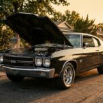 A dramatic, cinematic low-angle shot of a classic 1970 American muscle car — deep black with aggressive body lines and chrome accents — parked on a suburban driveway at golden hour, engine hood slightly propped, heat shimmer rising from the engine bay. The setting evokes a nostalgic 1970s American neighborhood with warm amber light casting long shadows across the asphalt. The car dominates the frame with a sense of raw, thunderous power held still. The atmosphere is reverent and emotional, like a memory frozen in time. Photorealistic style with cinematic depth of field, rich contrast, and a slightly faded vintage color grade that honors the era.