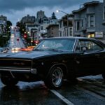A dramatic, cinematic photograph of a 1968 Dodge Charger R/T in glossy black, shot from a low three-quarter front angle on a rain-slicked urban street at dusk. The car's recessed tunneled grille, hidden headlights, and sweeping flying buttress C-pillars are sharply defined against a moody, deep blue-grey sky. Warm amber streetlights reflect off the sculpted body panels, highlighting the subtle body-side crease and the muscular fastback roofline. The four round taillights glow red at the rear. The setting evokes San Francisco's hilly streets, with blurred city lights in the background creating a sense of danger and motion even though the car sits still. Photorealistic, editorial automotive photography style with high contrast lighting, deep shadows, and a brooding, iconic atmosphere.