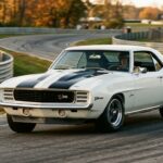 A dramatic, low-angle editorial photograph of a 1969 Chevrolet Camaro Z/28 on a winding road course, captured mid-corner with subtle tire lean suggesting precise, controlled handling. The car features its iconic hood stripe and front spoiler, rendered in classic white with bold racing stripes. The setting is golden hour, with warm light raking across the muscular, chiseled body lines of the restyled 1969 Camaro shape, emphasizing the pronounced hood and aggressive front end. In the background, a blurred track environment with armco barriers and autumn trees suggests a Trans-Am racing circuit. The overall mood is purposeful and driver-focused — not brute force, but mechanical precision and athletic balance. Shot in a cinematic, period-authentic style reminiscent of late 1960s motorsport photography, with slightly warm film-like tones and shallow depth of field that keeps the car sharp against a softly defocused background.
