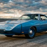 A dramatic, low-angle photograph of a 1969 Dodge Charger Daytona in vibrant corporate blue, shot on an open asphalt surface under a wide, cloud-streaked sky. The iconic 18-inch steel nose cone dominates the foreground, pointing aggressively toward the viewer, while the towering twin-post rear wing rises nearly two feet above the decklid in the background, silhouetted against the sky. The car's muscular, wide-body stance sits low and purposeful, with the pop-up headlights integrated cleanly into the aerodynamic nose. Harsh directional sunlight rakes across the body panels, emphasizing the sculptural curves and the sheer visual drama of the aerodynamic bodywork. The setting feels like an empty proving ground or superspeedway infield, evoking raw speed and engineering ambition. Photorealistic editorial automotive photography style, cinematic and high contrast, no text or logos visible.
