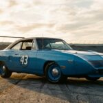 A dramatic low-angle photograph of a 1970 Plymouth Superbird in Petty blue, parked on sun-bleached asphalt at a wide oval superspeedway, the towering rear wing silhouetted against a vast open sky. The elongated nose cone stretches forward with purpose, the flush rear window gleaming, and every aerodynamic curve of the car catches harsh afternoon light. The composition emphasizes the car's extraordinary proportions — the improbable height of the wing, the aggressive forward thrust of the extended nose — making the machine look simultaneously alien and purposeful. Shot in a gritty, high-contrast editorial automotive photography style, with shallow depth of field, warm golden-hour lighting raking across the bodywork to highlight the sculpted surfaces, and the banked track curving away into the background, evoking the raw power and singular intensity of 1970s American motorsport.