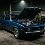 A dramatic, moody studio-style photograph of a 1969 Chevrolet Camaro ZL1 in a dark industrial setting, lit with a single dramatic overhead spotlight that casts deep shadows across the muscular body lines. The car is finished in a bold period-correct color, its aggressive front fascia and hood lines emphasized by the harsh directional lighting. The engine bay is open, revealing a gleaming all-aluminum 427 cubic inch big-block engine, its polished aluminum block and heads catching the light in stark contrast to the surrounding darkness. The atmosphere is raw and mechanical, with a concrete floor and faint traces of tire marks suggesting a drag strip or private warehouse. The composition is low and slightly angled, emphasizing the car's wide stance and purposeful aggression. The overall mood is secretive and legendary, evoking the sense of a rare, almost forbidden machine built outside the rules, a physical artifact of backroom ambition and racing obsession. Photorealistic, editorial automotive photography style with cinematic lighting.